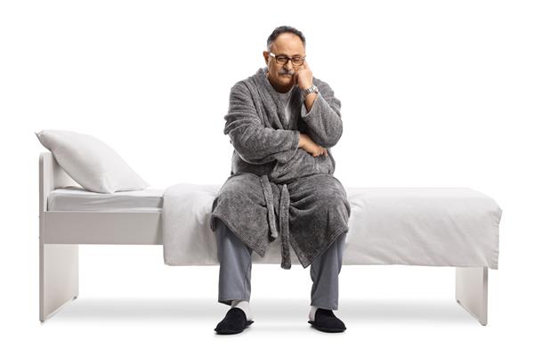 مرد بالغ متفکر و غمگین با لباس خواب و عبایی که روی تختی جدا شده در زمینه سفید نشسته است