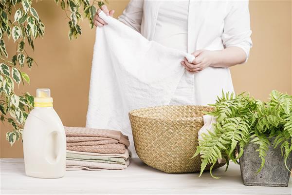 زن حوله سفید را در خانه تا می کند مجموعه ای از لباس های رنگی یک سبد لباسشویی بطری های مواد شوینده لباسشویی نرم کننده پارچه و یک گیاه آپارتمانی روی میز