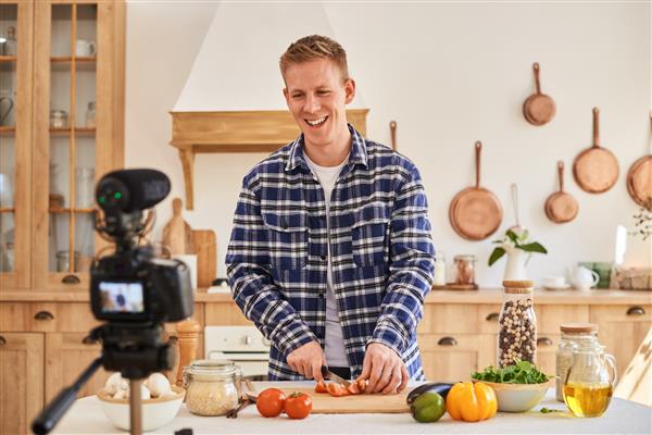 یک وبلاگ نویس مواد غذایی که محتوا تولید می کند و به دوربین نگاه می کند یک سرآشپز مرد خندان سبزیجات را برای سالاد برش می دهد و با دوربین روی سه پایه فیلم می گیرد تاثیر شبکه های اجتماعی