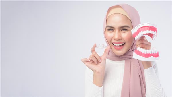 یک زن مسلمان جوان که بریس‌های اینویزیلاین و یک مدل دندان مصنوعی انسان را روی استودیو پس‌زمینه سفید مراقبت‌های بهداشتی دندان و مفهوم ارتودنسی در دست دارد