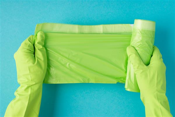 عکس نمای بالا از دست‌هایی با دستکش‌های لاستیکی سبز که کیسه‌های زباله سبز را در پس‌زمینه آبی پاستلی جدا شده با فضای کپی گرفته‌اند