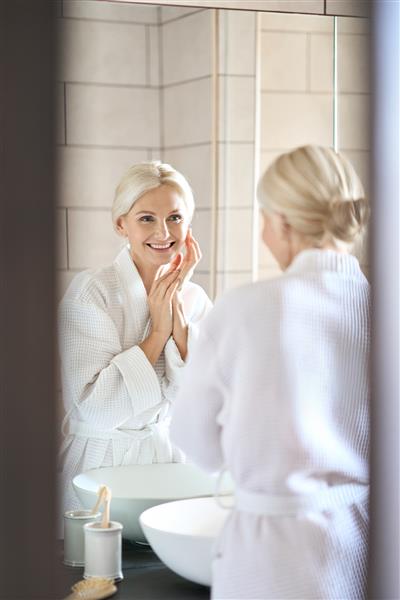 عکس عمودی از زن بلوند سالخورده جذاب دهه 50 در حمام با لباس حمام در حال لمس صورت پس از شستشو و زیبایی صبحگاهی که به انعکاس در آینه نگاه می کند و لبخند می زند مفهوم مراقبت از پوست
