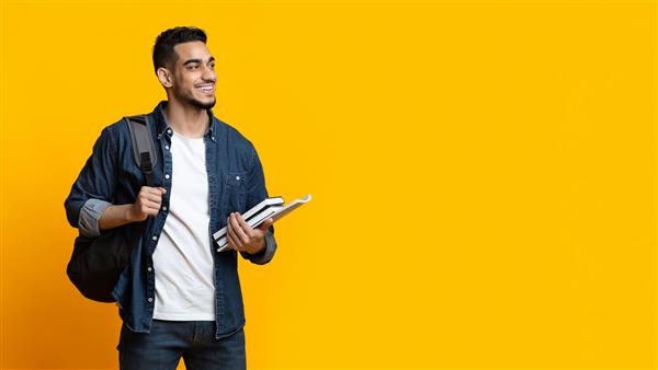 دانشجوی پسر مثبت عرب با کوله پشتی و دسته ای کتاب که به فضای کپی برای تبلیغات روی پس زمینه زرد استودیو پانوراما نگاه می کند آموزش دانشگاه کالج تحصیل مفهوم دوره