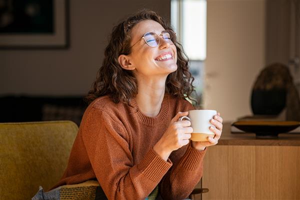 پرتره زن جوان شاد در حال لذت بردن از یک فنجان قهوه در خانه دختر زیبای خندان در حال نوشیدن چای داغ در زمستان زنی هیجان زده با عینک و ژاکت و خندیدن در یک روز پاییزی