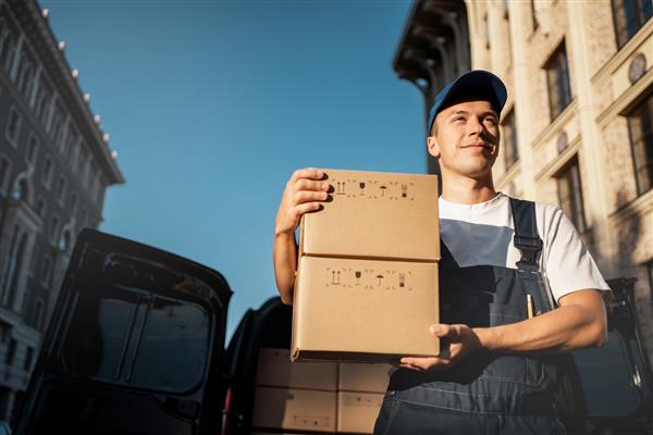 مردی با یونیفرم راحت مخصوص یک کارمند جعبه های مقوایی با تحویل درب منزل خدمات اکسپرس پیک لجستیک برای تحویل کالا از فروشگاه اینترنتی