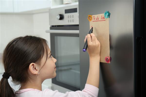 دختر کوچکی که در آشپزخانه لیست کارهایی که باید انجام شود روی یخچال می نویسد