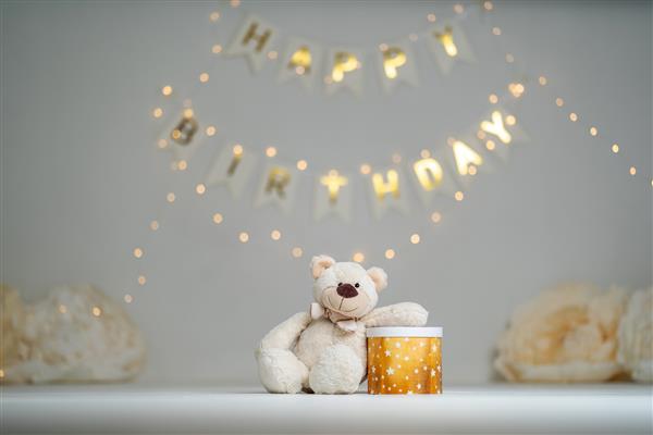 خرس عروسکی با جعبه هدیه روی جواهرات کودکانه با چراغ‌های درخشان گلدسته تولد کرم و گل‌های صورتی بزرگ می‌نشیند جشن اولین سال زندگی کودک منطقه عکس تزئین شده