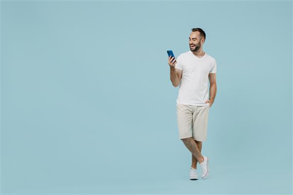 مرد جوان 20 ساله شاد و دوستانه خندان تمام قد با دوربینی با تی شرت سفید معمولی با استفاده از چت تلفن همراه onine جدا شده بر روی پرتره استودیویی پس‌زمینه آبی روشن پاستلی ساده
