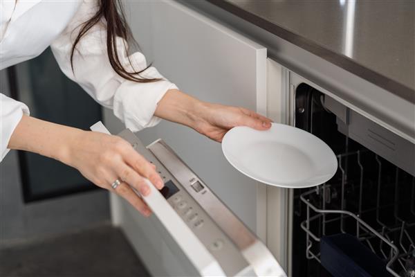 عکس برش خورده زن با استفاده از ماشین ظرفشویی توکار مدرن در حالی که مشغول انجام کارهای خانه در آشپزخانه در خانه است و بشقاب سفید داخل آن قرار داده است