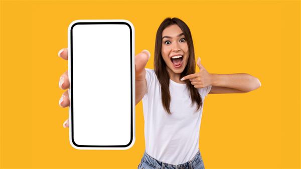 پیشنهاد عالی موبایل بانوی هیجان‌زده که با انگشت به گوشی هوشمند در دستش اشاره می‌کند واکنش عاطفی به اپلیکیشن جدید نشان می‌دهد زن هزاره‌ای شادمانه ایستاده روی پس‌زمینه استودیو نارنجی پانوراما