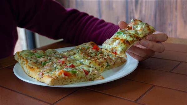دست زن در حال گرفتن یک تکه مستطیل شکل پیتزا مربع با ژامبون فلفل سبز گوجه فرنگی زیتون و پنیر از یک بشقاب سفید روی میز چوبی