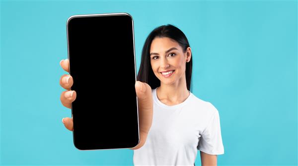 ماکت اپلیکیشن موبایل زن جوان زیبا در حال نمایش تلفن هوشمند بزرگ با صفحه سیاه سیاه زن خندان که فضای کپی رایگان را برای طرح یا تبلیغات شما نشان می دهد ژست روی پس زمینه آبی
