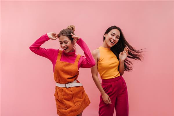 دو دختر جوان پرانرژی نژادی که در استودیو کنار هم می رقصند و دستانشان را تکان می دهند مدل‌هایی با موهای قهوه‌ای روشن و مشکی در لباس‌های بهاری نارنجی و صورتی روشن با لبخندی بر لب به سمت موسیقی حرکت می‌کنند