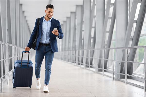 ورود آنلاین مرد خوش‌تیپ عرب که با چمدان در فرودگاه راه می‌رود و از گوشی هوشمند استفاده می‌کند پسر جوان خاورمیانه‌ای خندان در حال مرور اینترنت با تلفن همراه در حالی که به سمت دروازه پرواز می‌رود فضای کپی