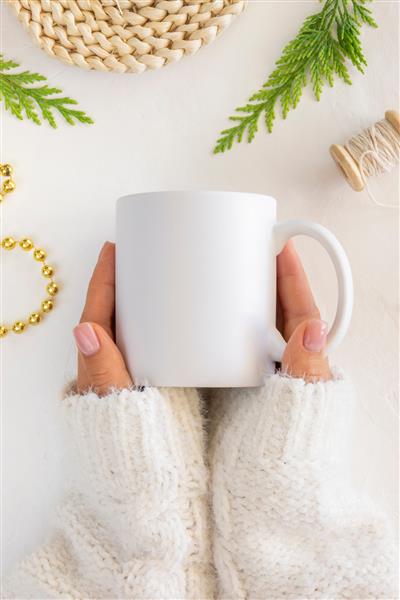 ماکت قهوه سفید از نزدیک دست های زنانه لیوان سرامیکی را نگه می دارد تخت دراز کشیده با شاخه درخت کریسمس و تزئینات کریسمس