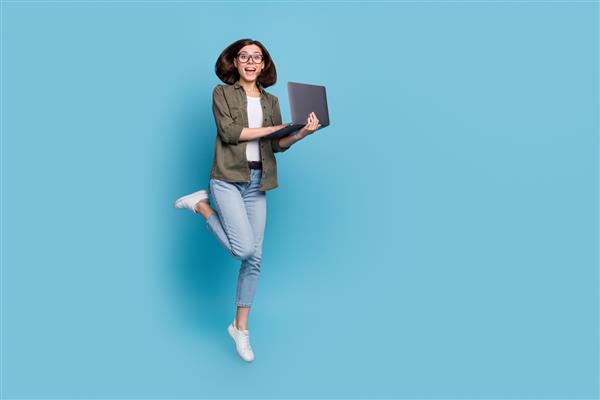 عکس تمام قد از بانوی هیجان زده کامپیوتری که توسعه دهنده پرش با استفاده از گجت استفاده می کند پیراهن شلوار جین ایزوله شده روی پس زمینه آبی رنگ