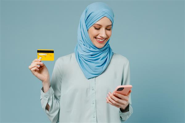 زن جوان مسلمان عرب آسیایی با حجاب عبایی تلفن همراه را با کارت بانکی اعتباری خرید آنلاین تحویل سفارش جدا شده در پس‌زمینه آبی ساده مردم شرق میانه اسلام مفهوم دینی