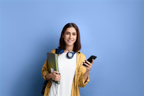 دختر دانشجوی خندان تلفن همراه در دست دارد به دوربین نگاه می کند از طریق برنامه آنلاین یاد می گیرد کتاب در دست دارد پیراهن زرد تی شرت سفید کیف مشکی و هدفون روی گردن دارد