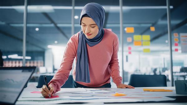 دفتر مدرن تاجر مسلمان با انگیزه با حجاب روی پروژه مهندسی کار می کند اسناد و نقشه ها را تجزیه و تحلیل می کند کارآفرین دیجیتال توانمند روی پروژه راه اندازی تجارت الکترونیک کار می کند