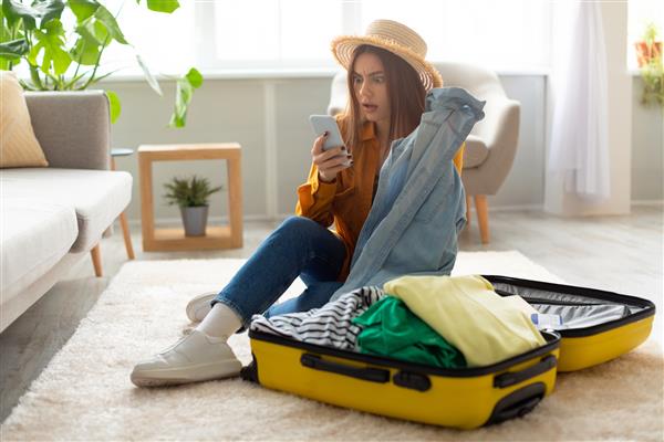 زن جوان وحشت زده در حال خواندن اخبار تکان دهنده سفر یا پیام منفی درباره گردشگری در گوشی هوشمند در حالی که چمدان خود را در خانه بسته است لغو پرواز مشکلات سفر در طول همه گیری کووید