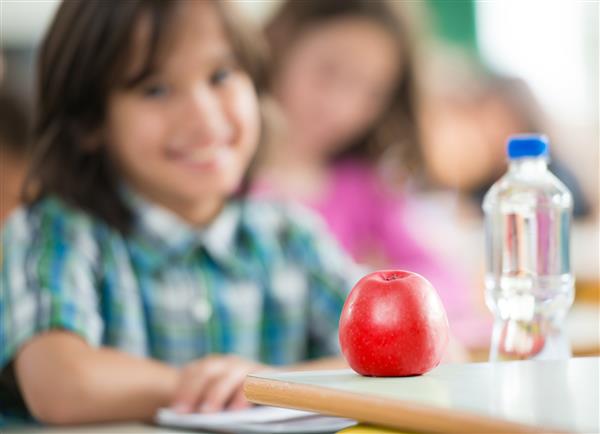 پسر کوچک شاد با بطری سیب و آب که در کلاس درس نشسته و لبخند می زند