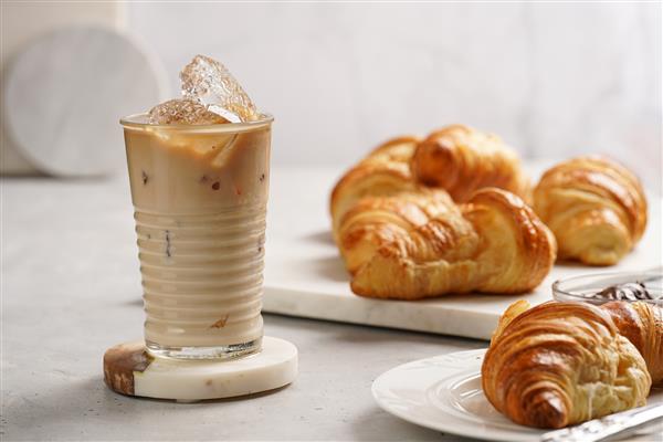 یک لیوان با قهوه شیر و تکه‌های یخ و کروسان‌های تازه پخته شده روی بشقاب مرمری کف‌پوش فندقی قهوه‌ای