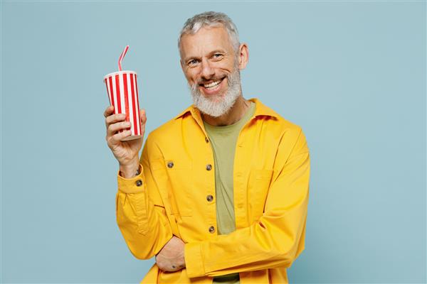 مرد سالخورده سبیل‌دار با موهای خاکستری شاد 50 ساله پیراهن زرد می‌پوشد فنجانی از نوشیدنی آب گازدار نوشابه‌کولا جدا شده بر روی پرتره استودیویی پس‌زمینه آبی روشن پاستلی ساده می‌پوشد مفهوم سبک زندگی مردم