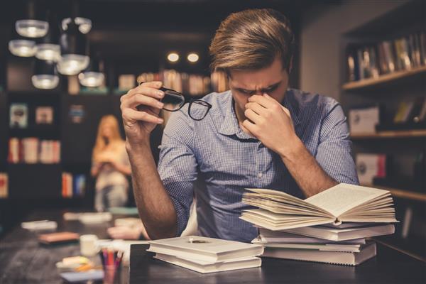 مرد جوان خسته در حالی که در کتابخانه مدرن به سختی کار می کند عینک در دست دارد و پل بینی خود را ماساژ می دهد