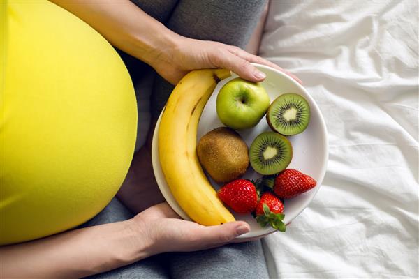 دختر حامله ای که یک کاسه میوه در دست دارد و روی یک تخت سفید نشسته است