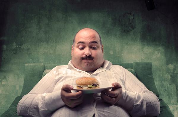 مرد چاق روی صندلی راحتی نشسته و به همبرگرش نگاه می کند