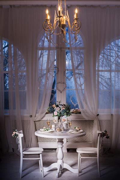 طراحی داخلی اتاق شب سفید با لوستر زیبا زیر میز سرو شده با گل روی آن پنجره های بزرگ در پس زمینه عمودی