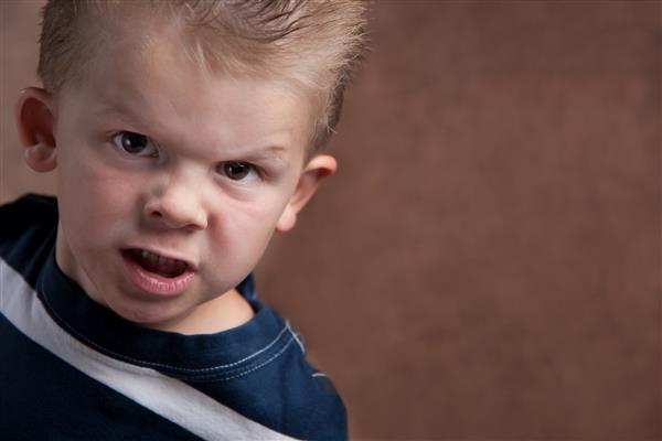 پسر کوچولوی عصبانی که روی پس‌زمینه بافت‌دار قهوه‌ای به دوربین خیره شده است