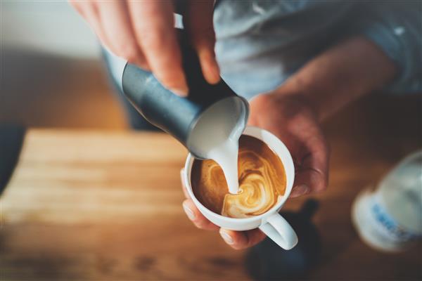 تصویر نزدیک از دستان مرد در حال ریختن شیر و تهیه کاپوچینوی تازه هنرمند قهوه و مفهوم آماده سازی قهوه صبح