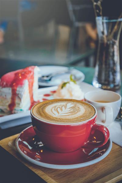 قهوه آروماتیک فنجان قرمز لاته آرت و کیک خوشمزه روی میز چوبی در کافی شاپ با رنگ تیره قدیمی و فضای کپی