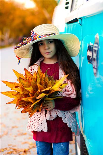 دختر بچه زیبا با کلاه و برگ های پاییزی زرد در دست