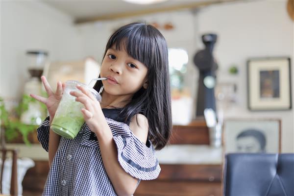 پرتره دختر بچه آسیایی تایلندی 4 تا 6 ساله یک کودک زیبا و زیبا با موهای بلند است یک فنجان چای سبز ماچا در دست دارد او دوست دارد برای رفع تشنگی سبزی بخورد او به یک کافه رفت