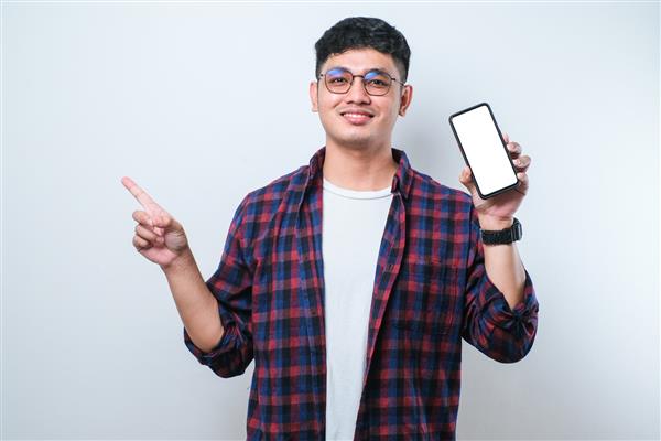 مرد جوان خوش‌تیپ آسیایی که صفحه خالی گوشی هوشمند را روی پس‌زمینه سفید جدا شده نشان می‌دهد بسیار خوشحال است که با دست و انگشت به پهلو اشاره می‌کند