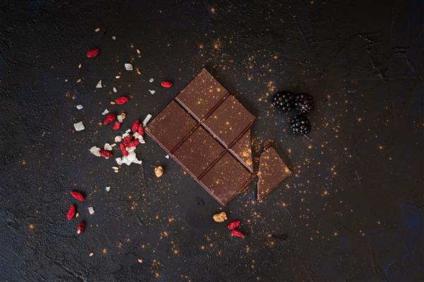 بلوک سیاه شکلاتی زیستی ارگانیک خوشمزه روی میز آبی تیره با بافت احاطه شده با آکای چیپس کوکو توت سیاه و پودر کاکائو مواد لذیذ