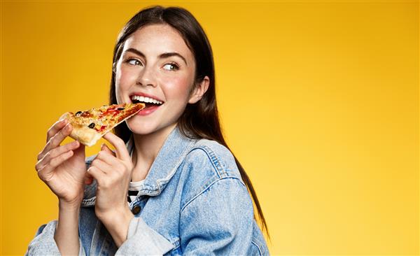 عکس زن جذابی که تکه‌ای از پیتزا را می‌خورد با انگشت شست کنار لباس‌های شیک پوشیده نشان می‌دهد که پیتزافروشی کجاست جدا شده روی پس‌زمینه زرد دختر زیبا میان وعده با فست فود می خورد