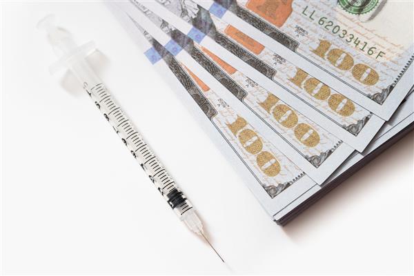 تصویر نزدیک از یک سرنگ پلاستیکی خالی که در نزدیکی یک پشته پول قرار دارد مفهوم هزینه‌های پیامدهای کووید19 و واکسیناسیون