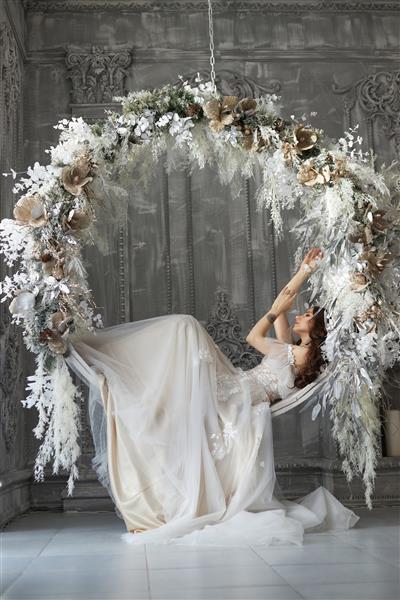 عروس زن با لباس سفید روی یک تاب گرد ساخته شده از گل های سفید و پرها تاب می خورد تصویری عاشقانه از زنی با خالکوبی روی بازویش عروسی