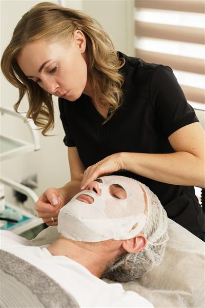 خانم متخصص زیبایی در حال زدن ماسک بر روی صورت مراجعه کننده در کلینیک زیبایی