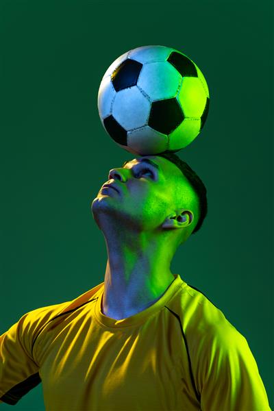 پرتره مرد جوان فوتبالیست با توپ روی سر که روی پس‌زمینه سبز استودیو در نور نئون ژست گرفته است