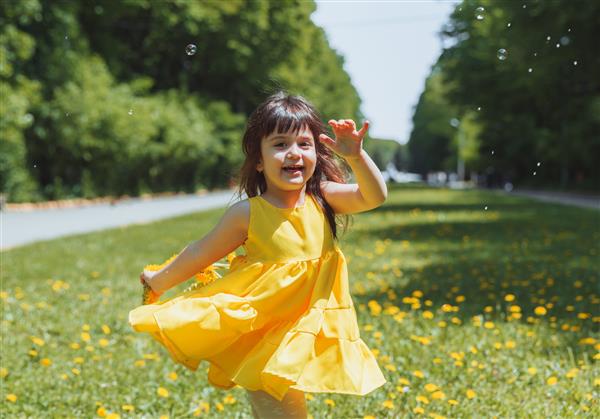 دختر بچه‌ای با لباس زرد تابستانی حباب‌های صابون را روی چمن‌های تاج گل پارک قاصدک‌ها می‌گیرد