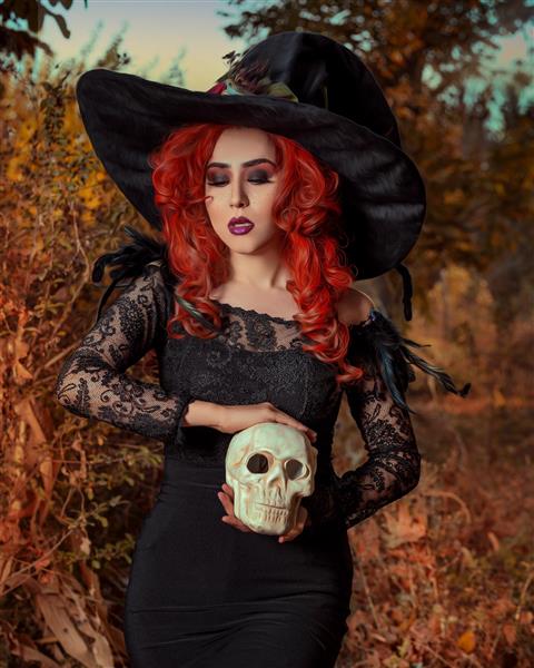 مدل زن با موهای قرمز و پیراهن سیاه و کلاه جادوگری و جمجمعه ای در دستانش