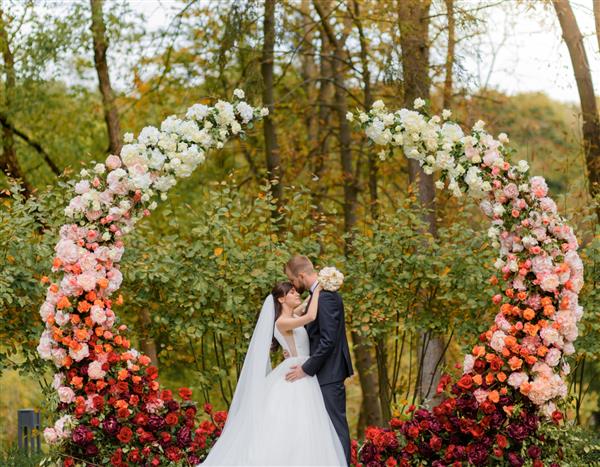 نمای جلوی تازه ازدواج کرده در یک مراسم عروسی در پارک در پس زمینه طاق گل