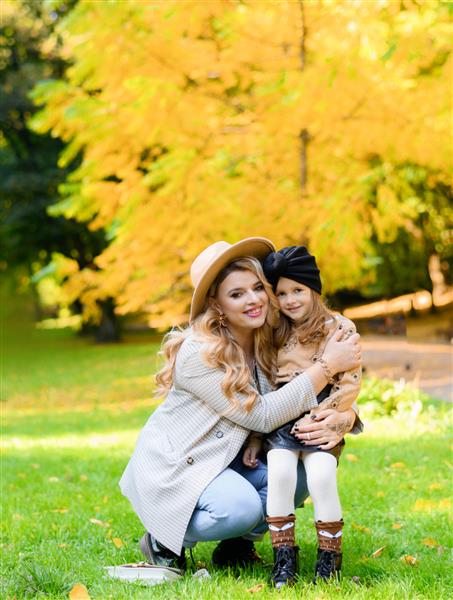 نمای روبروی مادر شاد با دختر دوست داشتنی با لباس های غیررسمی و کلاه در آغوش گرفتن و نشستن با هم در میان چمن ها در پس زمینه