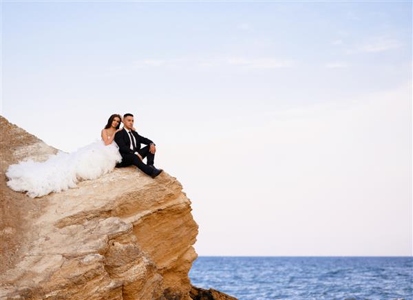 نمای جلوی زوج عروس جذاب که روی صخره نشسته اند و به چشم انداز دریا نگاه می کنند زن زیبا با لباس پف بلند و داماد دوست داشتنی را در آغوش گرفته و از طبیعت لذت می برند