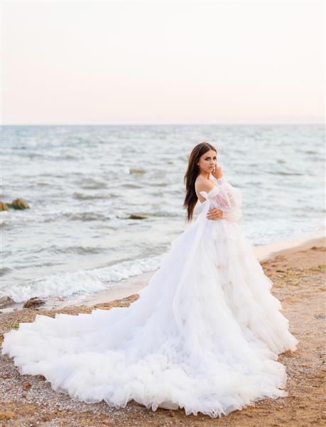 مدل زن عروس جذاب ایستاده در ساحل دریا با لباس عروس پف کرده زیبا که دست در دست گرفته نزدیک صورت جذاب منظره طبیعت در پس زمینه