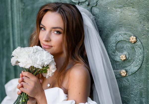نمای نزدیک از دختر جوان شایان ستایش با آرایش حرفه ای با لباس سفید با شانه های برهنه که دسته گل عروسی از گل های سفید را در نزدیکی صورت گرفته و در حالی که به در تکیه داده به دوربین نگاه می کند